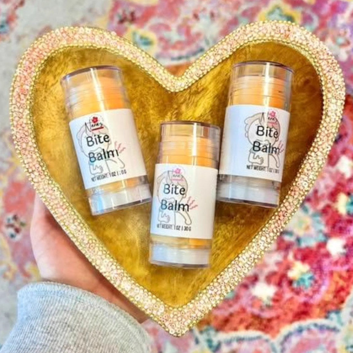Three 'Bite Balm' containers held in a heart-shaped gold holder against a colorful patterned background.