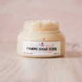 Jar of foaming sugar scrub with a visible brand label on a wooden surface.