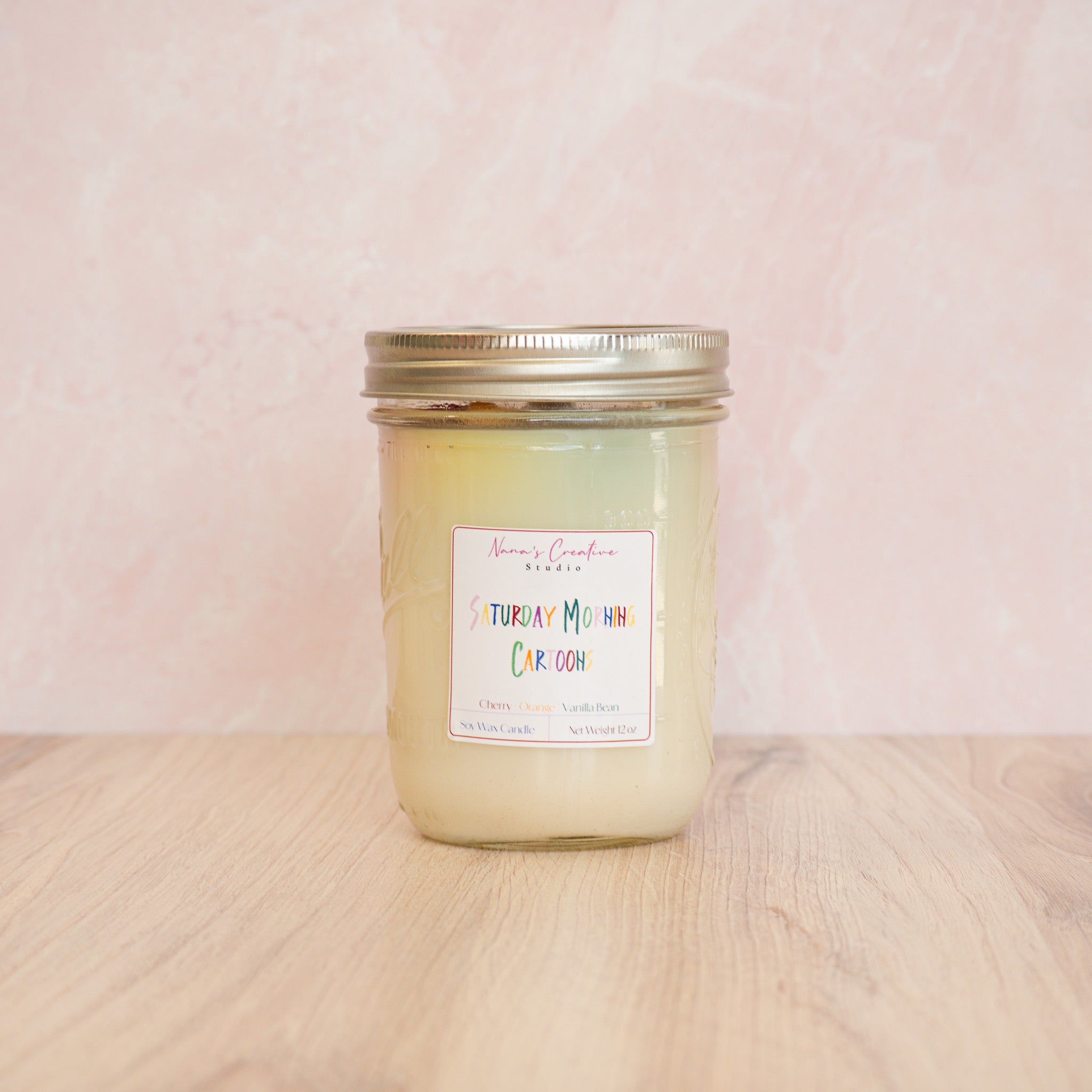 White candle in a jar labeled 'Saturday Morning Cartoons' on a white surface.